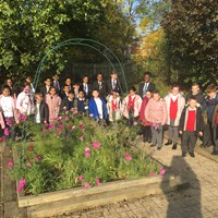 A large group of primary school children and two teachers stand around a large raised flowerbed with pink flowers.