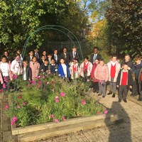 A large group of primary school children and two teachers stand around a large raised flowerbed with pink flowers.